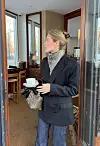 Person med svart blazer og skjerf bærer kaffekopp i døråpning til kafé.