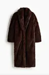 Lang mørkebrun fuskepelsjakke fotografert mot hvit bakgrunn.
