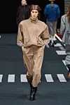 Modell i beige, voluminøst antrekk og brunt hodeplagg går nedover catwalk.
