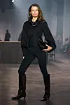 Modell på catwalken i mørkt antrekk med peplum-topp, bukser og høye støvler.