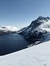 Snødekte fjell rundt en mørkeblå fjord under klar blå himmel