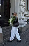 Kvinne i hvite ballongbukser og mørk jakke går i bygategate med blomsterbukett.