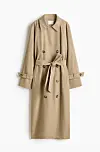 Beige, lang trenchcoat med belte og doble knapper mot hvit bakgrunn.