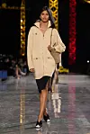Modell går på catwalk i beige jakke og svart skjørt foran opplyste kranstrukturer.