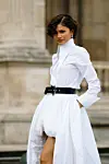 Person i hvit kjole med svart belte poserer utendørs foran klassisk bygning.