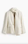 Kremfarget blazer med integrert skjerf hengt mot hvit bakgrunn.