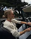 Kvinne i lys genser og solbriller kjører en åpen cabriolet på landevei.