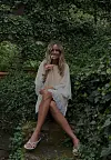 En kvinne sitter på en stol i en frodig hage med lys kjole og hvite sandaler.
