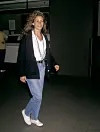 En kvinne med krøllete hår står innendørs i jeans, hvit topp, sort jakke og hvite sneakers.