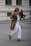 Kvinne i hvit bukse, trenchcoat, solbriller og svarte sandaler går over en gate.