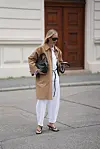En kvinne står i gaten iført beige trenchcoat, hvit skjorte, hvite jeans, svarte sandaler og en svart veske.