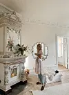 Lyst rom med dekorert antikk peis, stort speil, blomster og en kvinne som står på teppe ved vinduet.