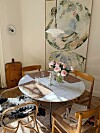 Lyst spisehjørne med rundt bord, blomster i vase, stoler, lysestaker og to store kunstverk på veggen.