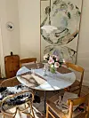 Lyst spisehjørne med rundt bord, blomster i vase, stoler, lysestaker og to store kunstverk på veggen.