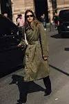 En kvinne i grønn trenchcoat og solbriller står ved en bil i en solfylt bygategate.