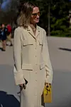 En person står utendørs i en lys beige lang kåpe og holder en gul veske.