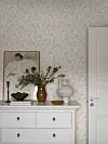 Lyst rom med blomstrete tapet, hvit kommode, speil, vase med greiner og en hvit dør.