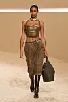 Modell går på en catwalk iført brun bermudashorts, kort topp, støvler og stor veske.