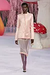 Modell på catwalk iført lys rosa jakke og knelangt plagg i et pastellfarget moteshow.
