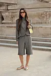 En kvinne står foran en steintrapp i grå blazer, bermudashorts, solbriller og loafers med en liten gul veske.