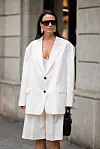 Kvinne i kremhvit blazer og knelange culottes står foran en bygning og holder en svart veske.