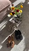 Tre par sko står på gulvet ved siden av en vase med fargerike blomster og noen magasiner.