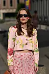 Kvinne i rosa blomsterdress, mørke solbriller og med veske står utendørs.