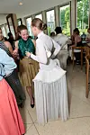 Gjester i sommerklær står og prater innendørs ved store vinduer med utsikt over byen.
