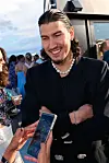 En smilende mann i svart dress står på en takterrasse mens noen holder en mobiltelefon foran ham.