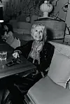 En smilende kvinne sitter i en sofa ved et bord i et loungeområde med dekorert vegg i bakgrunnen.
