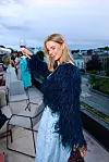 Kvinne i blått antrekk skåler med et vinglass på en takterrasse med utsikt over byen.