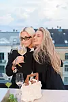 To personer står tett sammen på en takterrasse, den ene holder et vinglass foran en kake på bordet.
