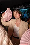En mann i beige dress holder en rosa solfjær på en fest med folk i bakgrunnen.