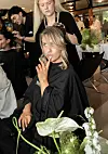En kvinne får håret stylet mens en makeupartist jobber i bakgrunnen i en lys lounge med speil og blomster.