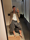 Person tar speilselfie i en beige blazer, hvit topp og mørk bermudashorts i en lys gang.