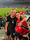To personer står tett sammen på tribunen på et fotballstadion, kledd i rødt og svart.