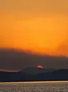 Solnedgang med rød sol bak mørke fjell og oransje himmel over et rolig hav.