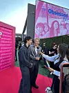 To menn står på rosa teppe ved en presseopptreden foran en stor «Obsessed»-vegg og blir intervjuet.