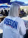 Person sett bakfra i hvit jakke med «Obsession»-tekst ved en uteservering med blå parasoller.
