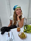 Kvinne med fargerikt hodeplagg sitter ved et bord med en svart hund, drikke og en skål med oliven.
