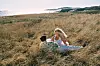 To personer i høyt gress ved kysten med havet i bakgrunnen.