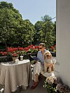 En kvinne sitter med en hund ved et lite bord på en terrasse med blomster og trær i bakgrunnen.
