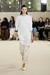 En modell går på en catwalk i en hvit, løs kjole og lyse bukser foran en sittende publikum.