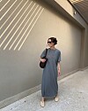 Kvinne i grå T-skjortekjole, solbriller og sandaler står ved en beige vegg med skygger.