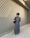 Kvinne i grå T-skjortekjole, solbriller og sandaler står ved en beige vegg med skygger.