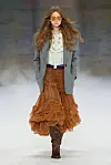 Modell på catwalken for Chloé i grå blazer, hvit bluse, rutete rustrødt skjørt og brune støvler.