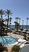 Bassengområde på Lucia Marbella med palmer, hvite parasoller og havet i bakgrunnen.