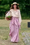 Modell i lys bluse, rosa skjørt og hatt går på en gresskledd catwalk med en kurv i hånden.
