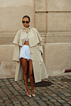 Person i lys trenchcoat, hvit shorts og solbriller står foran en beige steinvegg.