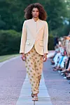 En modell går på en utendørs catwalk i en lys, krageløs blazer og et langt mønstret skjørt.